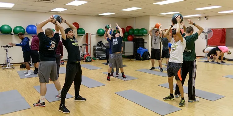 Students in a gym passing weighted balls to each other, with mats on the floor